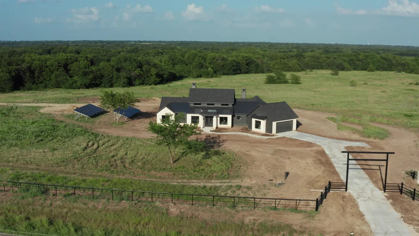 Aerial view of a new Ark Industries estate home with private drive, steel ranch gate, and pipe fencing on green acreage
