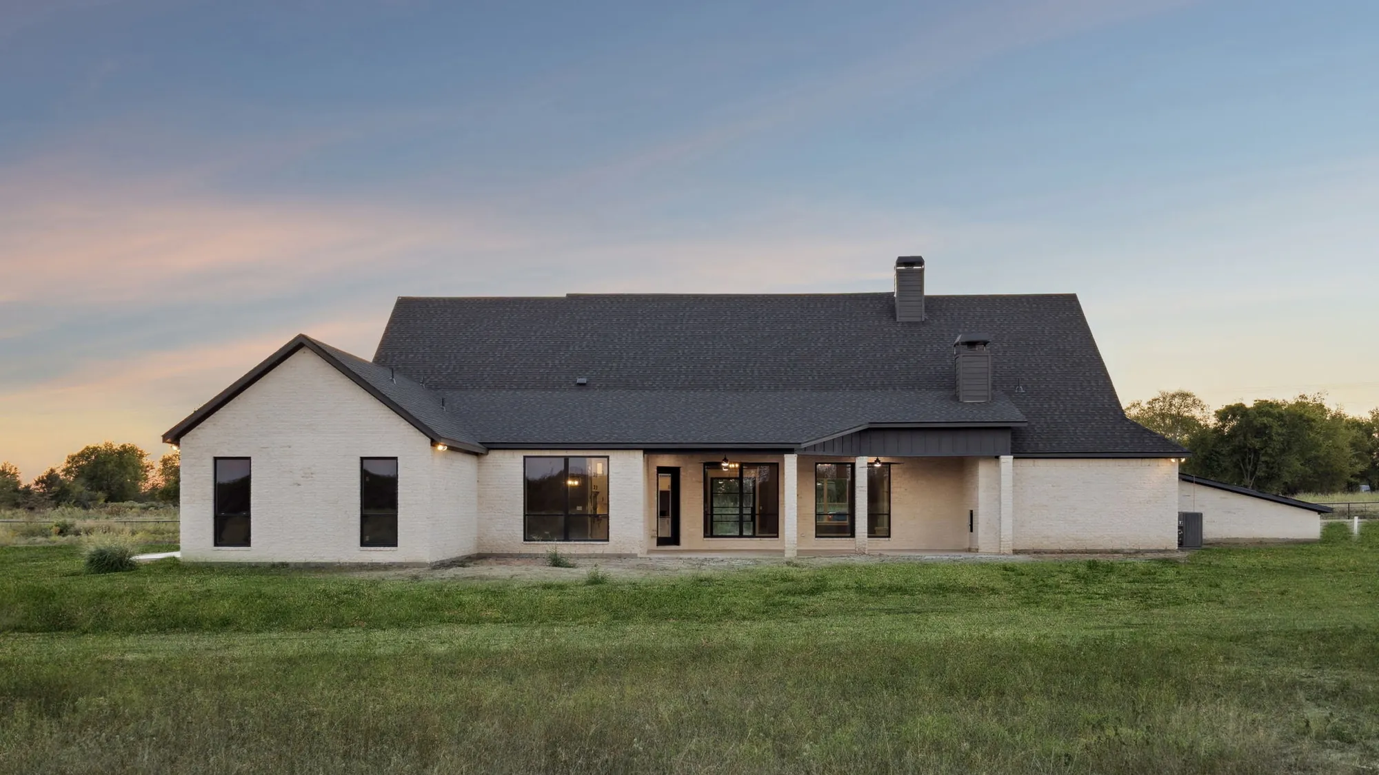 Rear elevation of a custom home at dusk, interior light glowing through tall windows onto the lawn